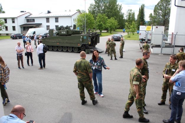 Soldiers and participants gathered outdoors at a military camp, an armoured vehicle in the background